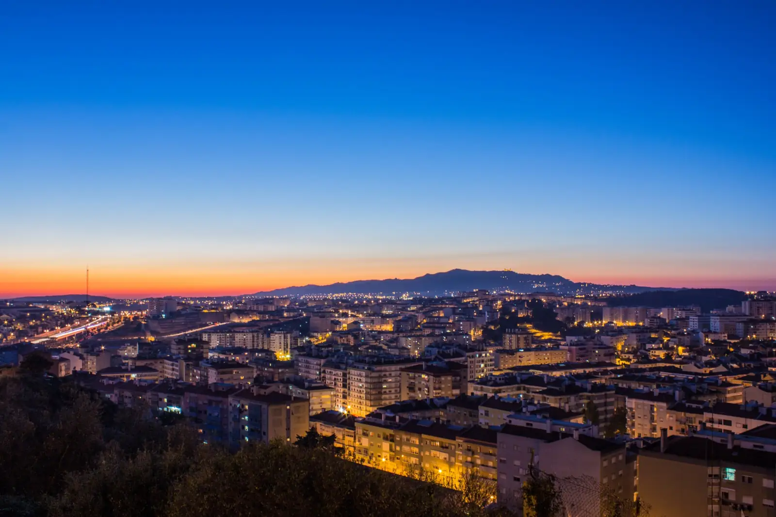 Agualva-Cacém ao entardecer, vista de cima: céu azul, prédios iluminados e, ao fundo, a Serra de Sintra.
