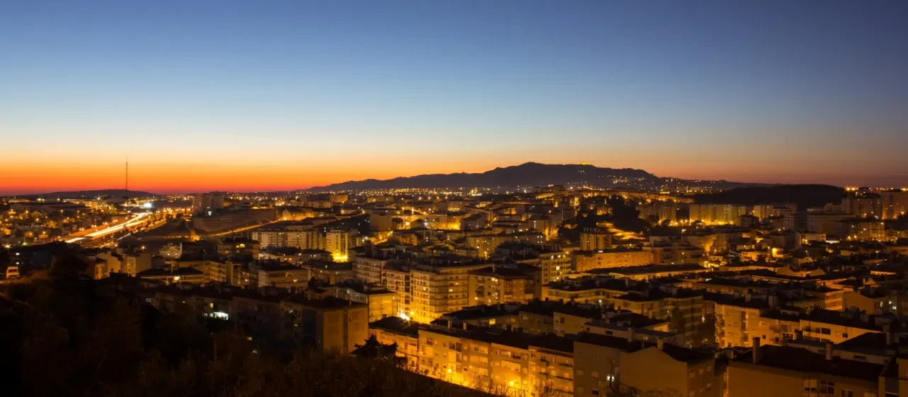 Vista real de Agualva-Cacém ao entardecer: prédios iluminados e, ao fundo, a Serra de Sintra.
